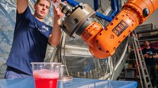 In einer großen Werkhalle demonstriert eine Person die Arbeit an einem orangefarbenen Industrieroboter, indem sie ein Bauteil einspannt. Im Vordergrund steht ein Plastikbecher mit rotem Getränk. Eine weitere Person beobachtet die Szene aus einiger Entfernung.