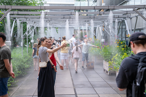 Viele Menschen gehen durch einen überdachten Außenbereich, in dem aus mehreren Düsen feiner Wassernebel versprüht wird. Einige Kinder spielen im Nebel, Erwachsene machen Fotos oder genießen die Abkühlung. Links und rechts stehen dichte Pflanzen und Blumenbeete.