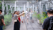 Viele Menschen gehen durch einen überdachten Außenbereich, in dem aus mehreren Düsen feiner Wassernebel versprüht wird. Einige Kinder spielen im Nebel, Erwachsene machen Fotos oder genießen die Abkühlung. Links und rechts stehen dichte Pflanzen und Blumenbeete.