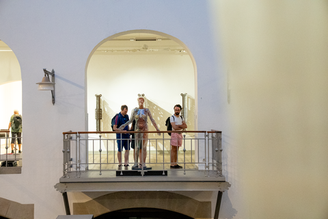 Auf einer Galerie innerhalb eines Museums stehen drei Personen neben einer lebensgroßen, transparenten Figur, die menschliche Anatomie zeigt. Hinter ihnen befinden sich hohe Rundbögen und weitere Ausstellungsstücke.