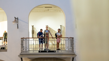Auf einer Galerie innerhalb eines Museums stehen drei Personen neben einer lebensgroßen, transparenten Figur, die menschliche Anatomie zeigt. Hinter ihnen befinden sich hohe Rundbögen und weitere Ausstellungsstücke.