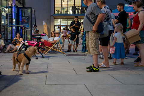 Vor einem Gebäude sitzen und stehen mehrere Menschen in entspannter Atmosphäre. Ein vierbeiniger Roboter in pink-weißer Farbgebung steht zwischen ihnen, während ein echter Hund neugierig vor ihm steht. Einige Menschen sitzen in Liegestühlen, andere beobachten die Szene oder machen Fotos.