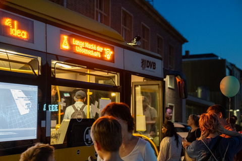 Bei Dunkelheit steht eine Menschengruppe vor einer gelben Straßenbahn der DVB, deren Display „Lange Nacht der Wissenschaft“ anzeigt. Im Inneren der Bahn laufen technische Vorführungen, die durch das Fenster sichtbar sind. Menschen unterhalten sich, ein Kind hält einen Luftballon.
