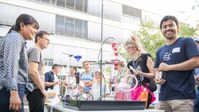 Draußen um einen Tisch mit Laboraufbau versammelt sich eine Gruppe von Erwachsenen und Kindern. Verschiedene Glasgefäße, Schläuche und farbige Flüssigkeiten bilden eine experimentelle Chemie‑Station. Die Stimmung ist fröhlich und neugierig.