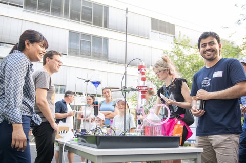 Draußen um einen Tisch mit Laboraufbau versammelt sich eine Gruppe von Erwachsenen und Kindern. Verschiedene Glasgefäße, Schläuche und farbige Flüssigkeiten bilden eine experimentelle Chemie‑Station. Die Stimmung ist fröhlich und neugierig.