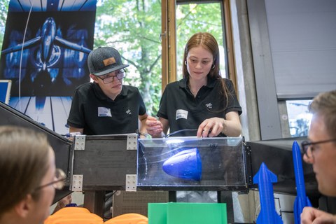 Zwei Personen mit dunklen Polohemden demonstrieren ein Experiment an einem Wasserkanalmodell. Sie halten ein blaues Objekt im strömenden Wasser, während mehrere Zuschauerinnen und Zuschauer gespannt zuschauen.