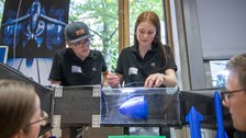 Zwei Personen mit dunklen Polohemden demonstrieren ein Experiment an einem Wasserkanalmodell. Sie halten ein blaues Objekt im strömenden Wasser, während mehrere Zuschauerinnen und Zuschauer gespannt zuschauen.