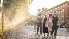 Eine Gruppe junger Menschen läuft lachend eine Kopfsteinpflasterstraße entlang. Links sprühen Rasensprenger feinen Wassernebel in die warme Abendsonne, wodurch die Umgebung golden schimmert.