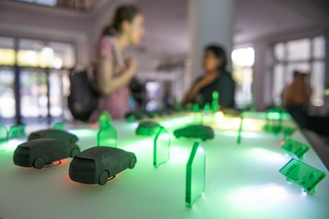 Auf einem Tisch stehen viele kleine Modellautos und Gebäudeteile aus grünlichem Acryl, die von unten farbig beleuchtet werden. Im Hintergrund unterhalten sich zwei Personen, die unscharf abgebildet sind.