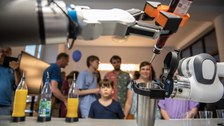 Zwei Roboterarme mixen ein Getränk: Einer hält einen Metallbecher, der andere gießt rote Flüssigkeit aus einer Flasche hinein. Auf dem Tisch stehen mehrere Saftflaschen mit Dosieraufsätzen. Im Hintergrund beobachten Erwachsene und Kinder interessiert die Demonstration.
