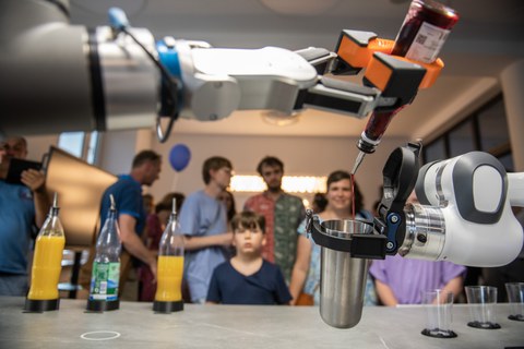 Zwei Roboterarme mixen ein Getränk: Einer hält einen Metallbecher, der andere gießt rote Flüssigkeit aus einer Flasche hinein. Auf dem Tisch stehen mehrere Saftflaschen mit Dosieraufsätzen. Im Hintergrund beobachten Erwachsene und Kinder interessiert die Demonstration.
