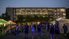 Am Abend schlendern viele Menschen über eine Wiese zwischen Zelten und bunten Lichtern. Im Hintergrund leuchtet eine modern verglaste Gebäudefassade, und die Leute wirken entspannt wie auf einem Sommerfest.