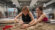 Zwei Personen beugen sich konzentriert über ein sandiges Wasserbecken in einer großen Versuchshalle. Mit den Händen formen sie Strukturen im nassen Sand, während im Hintergrund technische Anlagen und Laborgeräte zu sehen sind.