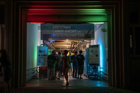 Ein Eingang ist bunt beleuchtet und mit einem Banner „Dresdner Lange Nacht der Wissenschaften“ versehen. Menschen gehen hindurch zu einer Ausstellung mit technischen Geräten und Informationspostern im Inneren.