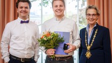 Drei Personen stehen zusammen und lächeln. Die Person in der Mitte hat einen Blumenstrauß, eine Mappe und ein kleines Etui in der Hand. Die Person rechts trägt erneut eine goldene Amtskette.