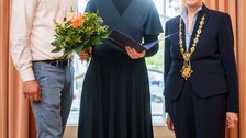 Drei Personen stehen in einem festlich wirkenden Raum vor einem großen Fenster. Die Person in der Mitte hält eine geöffnete Mappe mit einer Urkunde und lächelt in die Kamera. Die Person links hält einen Blumenstrauß, die Person rechts trägt eine goldene Amtskette.