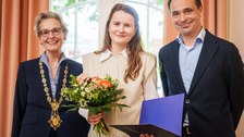 Drei Personen posieren gemeinsam vor einem großen Fenster. Die Person in der Mitte hält einen bunten Blumenstrauß sowie eine geöffnete Mappe. Die Person links trägt eine goldene Amtskette, die Person rechts steht lächelnd daneben.