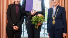 Drei Personen stehen nebeneinander vor einem großen Fenster. Die Person in der Mitte hält eine geöffnete Mappe mit einer Urkunde sowie einen Blumenstrauß. Die beiden Personen links und rechts flankieren die ausgezeichnete Person und lächeln in die Kamera; die Person rechts trägt eine goldene Amtskette.