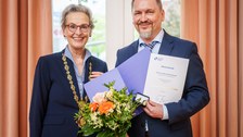 Zwei Personen stehen nebeneinander und halten gemeinsam eine Mappe mit einer Urkunde sowie einen Blumenstrauß. Beide lächeln in die Kamera; die Person links trägt eine goldene Amtskette.