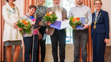 Sechs Personen stehen in einer Reihe vor einem großen Fenster. Mehrere von ihnen halten Blumensträuße und geöffnete Mappen mit Urkunden. Die Stimmung wirkt feierlich und wertschätzend.