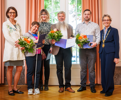 Sechs Personen stehen in einer Reihe vor einem großen Fenster. Mehrere von ihnen halten Blumensträuße und geöffnete Mappen mit Urkunden. Die Stimmung wirkt feierlich und wertschätzend.