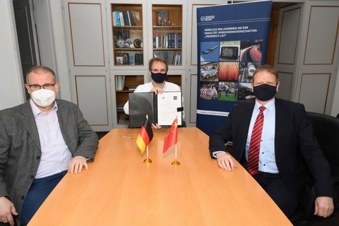 Drei Herren mit Maske sitzen an einem hellbraunen Tisch. Der Herr in der MItte hält eine Mappe in der Hand.