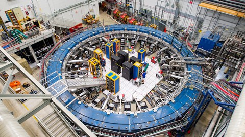 Blick auf den blauen Muon g-2 Ring, er befindet sich in seiner Detektorhalle inmitten von Elektronikregalen, der Myonen-Beamline und anderen Geräten.