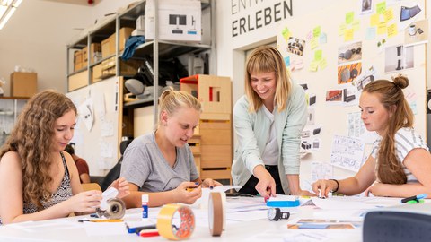 Drei junge Frauen sitzen an einem Tisch und basteln, eine Frau steht dazwischen und zeigt auf die Bastelei.