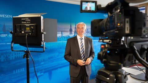 Prof. Dr. Jürgen Stamm führt durch die Online-Veranstaltung, rechts im Vordergrund ein Monitor, links eine Kamera, hinter Prof. Stamm ein blauer Hintergrund.