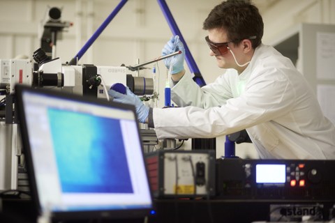 Eine Person im weißen Kittel steht rechts im Bild und bedient eine wissenschaftliche Apperatur. Im linken unteren Bildrand ist ein blaues Display zu sehen.
