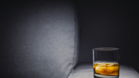 Foto von einem Glas Whiskey mit Eis, das auf einer grauen Sofalehne steht. 