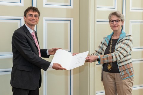 Prof. Friedrich (links) und Prof. Staudinger bei der Übergabe einer Urkunde, beide blicken in die Kamera.
