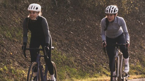 Zwei Frauen in Sportkleidung fahren im Freien Rennrad. Beide tragen einen Schutzhelm.