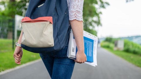 Eine Studentin spaziert am Blauen Wunder Dresden. Sie trägt einen blauen Rucksack auf dem Rücken und hält Hefter und Buch in der rechen Hand.