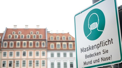 Vor dem Hintergrund einer Altbaufassade steht rechts ein Schild, das auf Maskenpflicht hinweist.