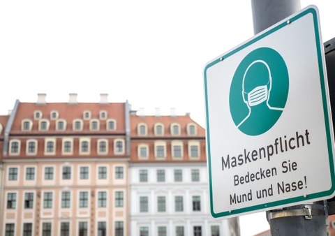 Vor dem Hintergrund einer Altbaufassade steht rechts ein Schild, das auf Maskenpflicht hinweist.
