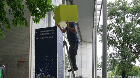 Eine Person steht auf einer Leiter vor dem Hörsaalzentrum und montiert einen hellgrünen Würfel auf den dunkelblauen Wegweiser.