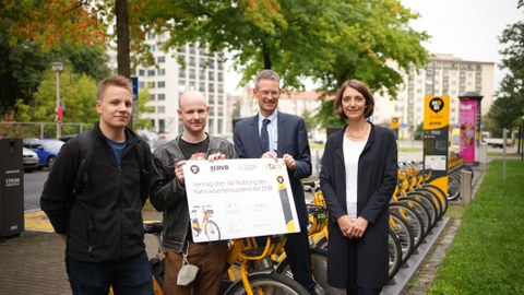 Vier Personen stehen im Halbkreis vor einer Reihe gelber Fahrräder. Die zweite Person von links hält einen symbolischen Vertrag in der Hand.