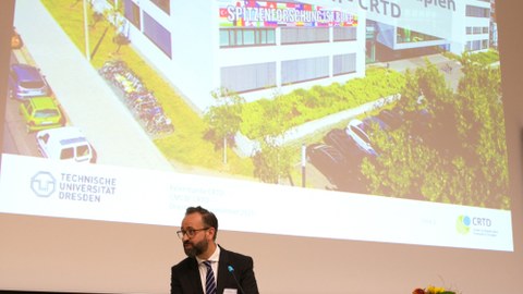 Wissenschaftsminister Sebastian Gemkow zur CRTD Feierstunde, im Hintergrund ein Foto des crtd-Gebäudes 	