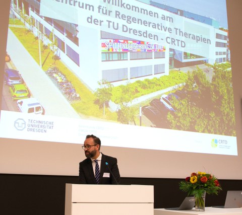 Wissenschaftsminister Sebastian Gemkow zur CRTD Feierstunde, im Hintergrund ein Foto des crtd-Gebäudes 	