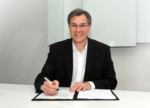 Porträtfoto Dr. Lamprecht vor einer geöffneten Unterschriftsmappe, in der rechten Hand hält er ein Schreibgerät.