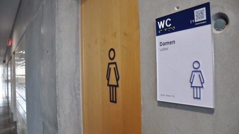 Picture of a women's restroom highlighting the sign written in English and German, featuring Braille, tactile lettering, symbols and a QR code.