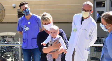 Eine blonde Frau mit Mundschutz hält ein Baby auf dem Arm, sie wird umringt von Pflegepersonal