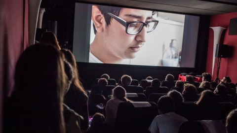 Blick in einen dunklen Kinosaal, auf der Leinwand ist ein junger Mann mit Brille zu sehen.