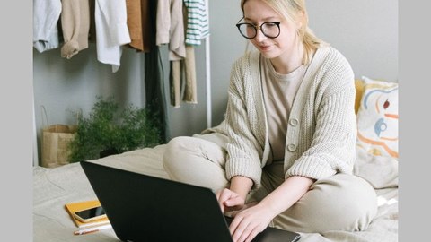 Eine junge, blonde Frau mit Zopf und Brille sitzt auf einem Bett und schaut in den aufgeklappten Laptop.