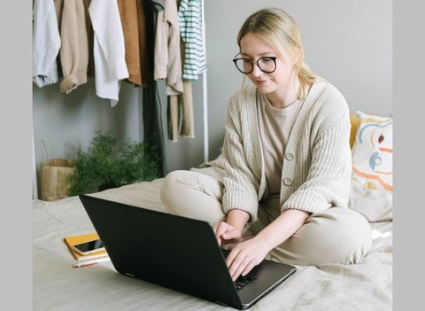 Eine junge, blonde Frau mit Zopf und Brille sitzt auf einem Bett und schaut in den aufgeklappten Laptop.