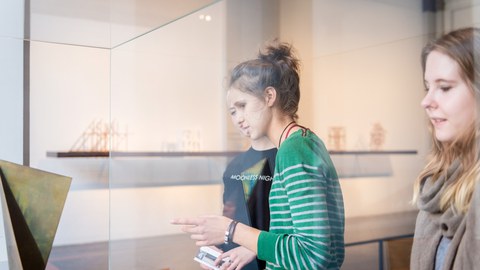 Foto mit drei jungen Frauen, die seitlich vor einer Vitrine stehen.