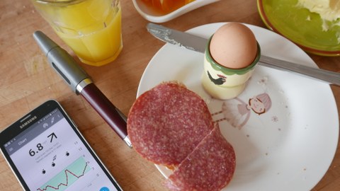 Auf einem weißen Frühstücksteller befinden ein Ei im Eierbecher und ein Salamibrot. Links daneben liegen ein Kuli und ein Smartphone, auf dem ein Diagramm zu sehen ist.