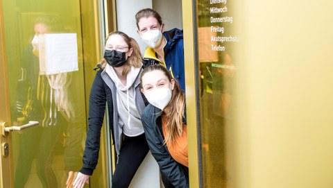 Drei junge Frauen mit Mundschutz schauen aus einer geöffneten gelben Glastür.