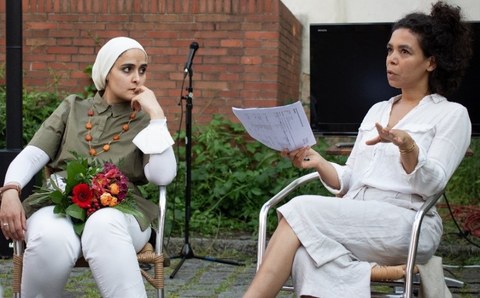 Links sitzt eine Frau mit weißem Kopftuch, einen Blumenstrauß auf dem Schoß, rechts eine Frau im weißen Kleid, sie spricht und hält einen Bogen Papier in der Hand. Im Hintergrund eine Backsteinwand und eine Hecke, davor ein Mikrophon.